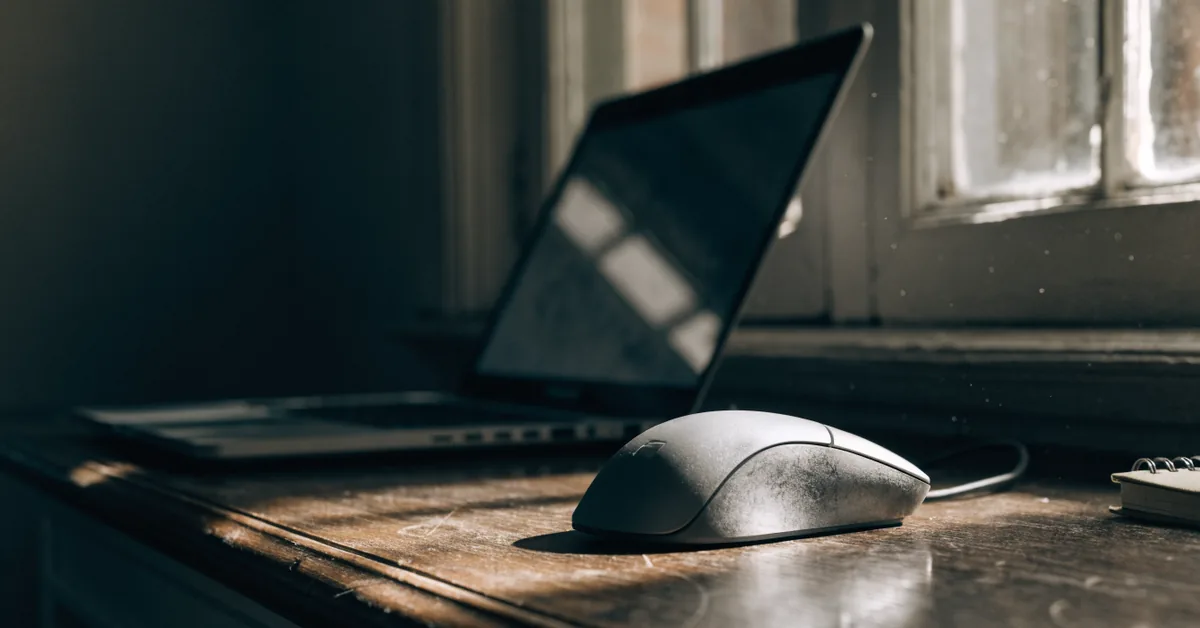 Computer mouse on a wooden desk in low window light, an open laptop behind it